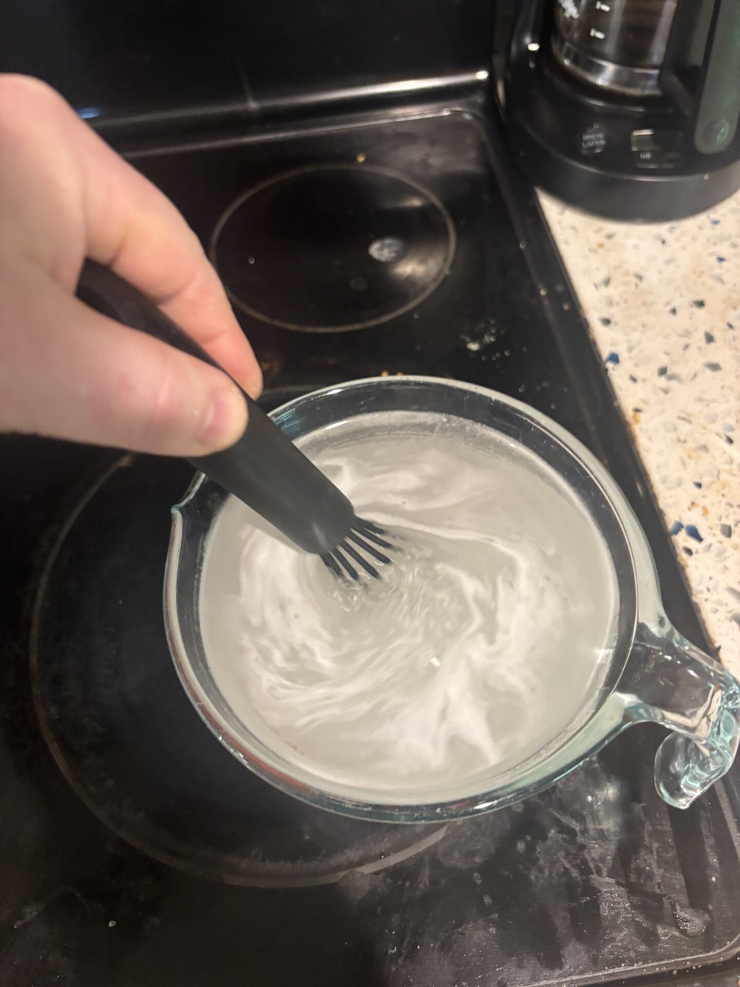 Sugar syrup in a glass measuring cup, mid-stir with a black silicone brush. White swirls dissolving into clear water.