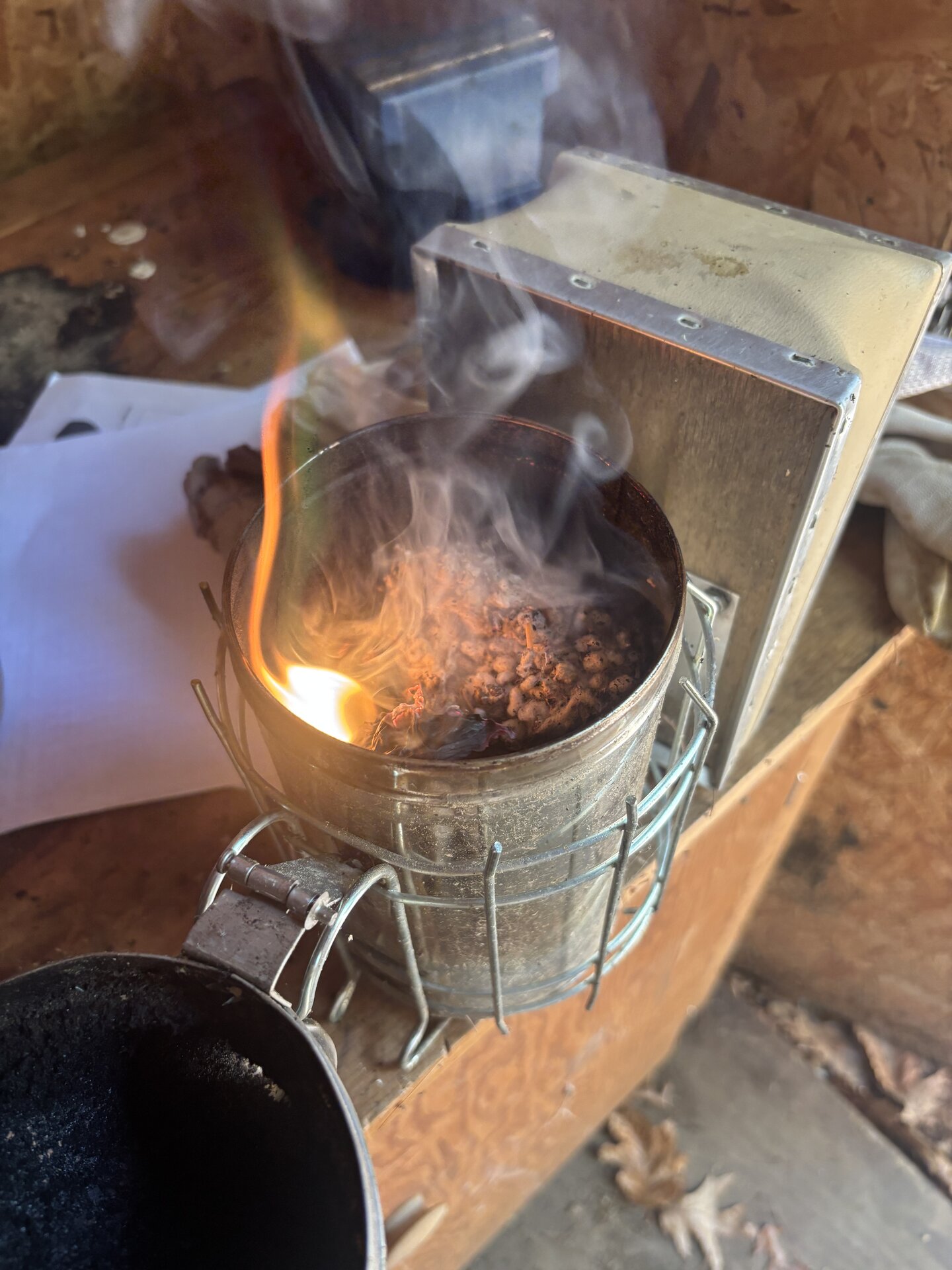 A metal bee smoker burning on a workbench. Orange flame and white smoke from a tin of smoldering pellets.